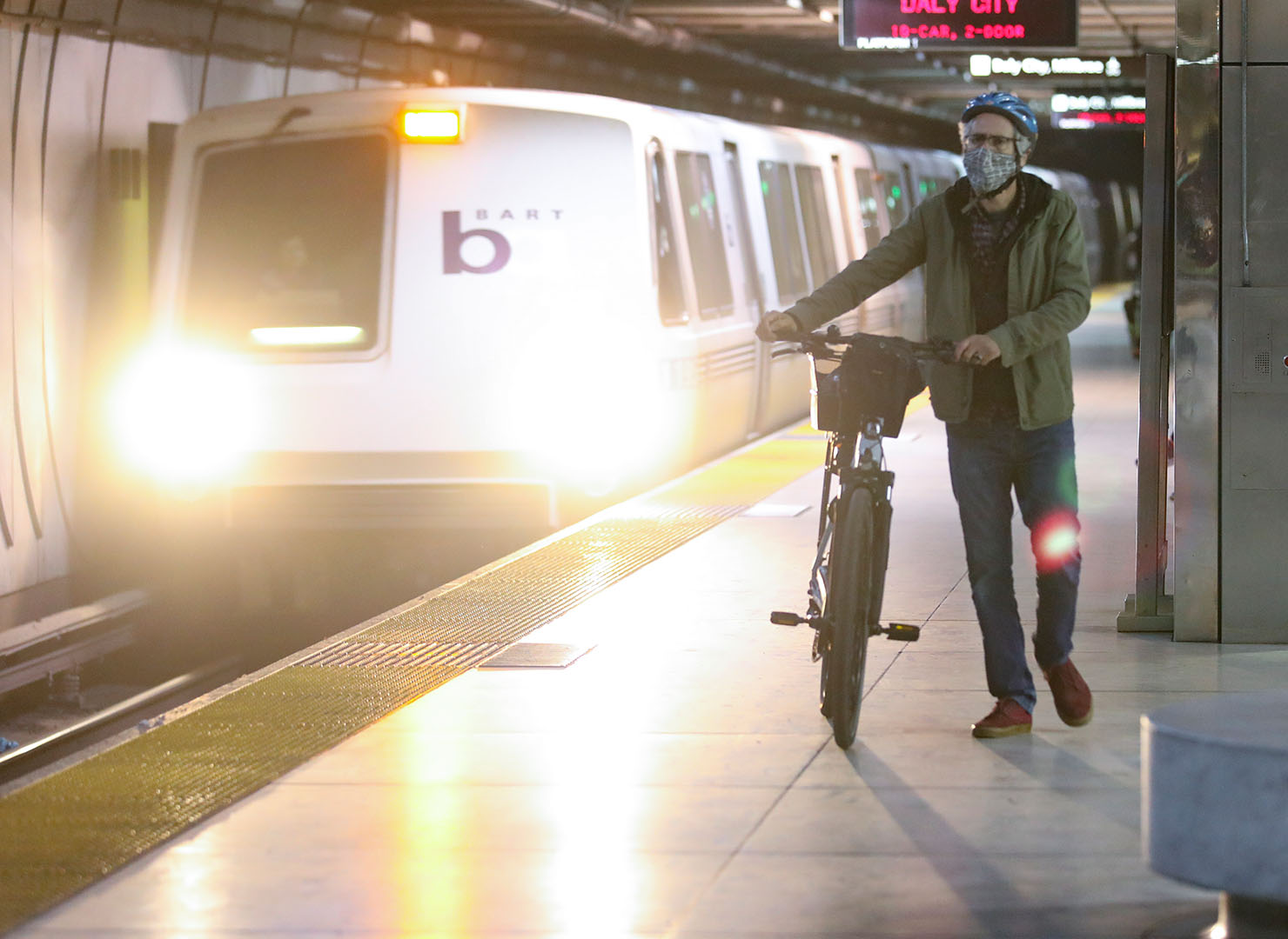 A masked cyclist walks with his bike through an underground BART station. 