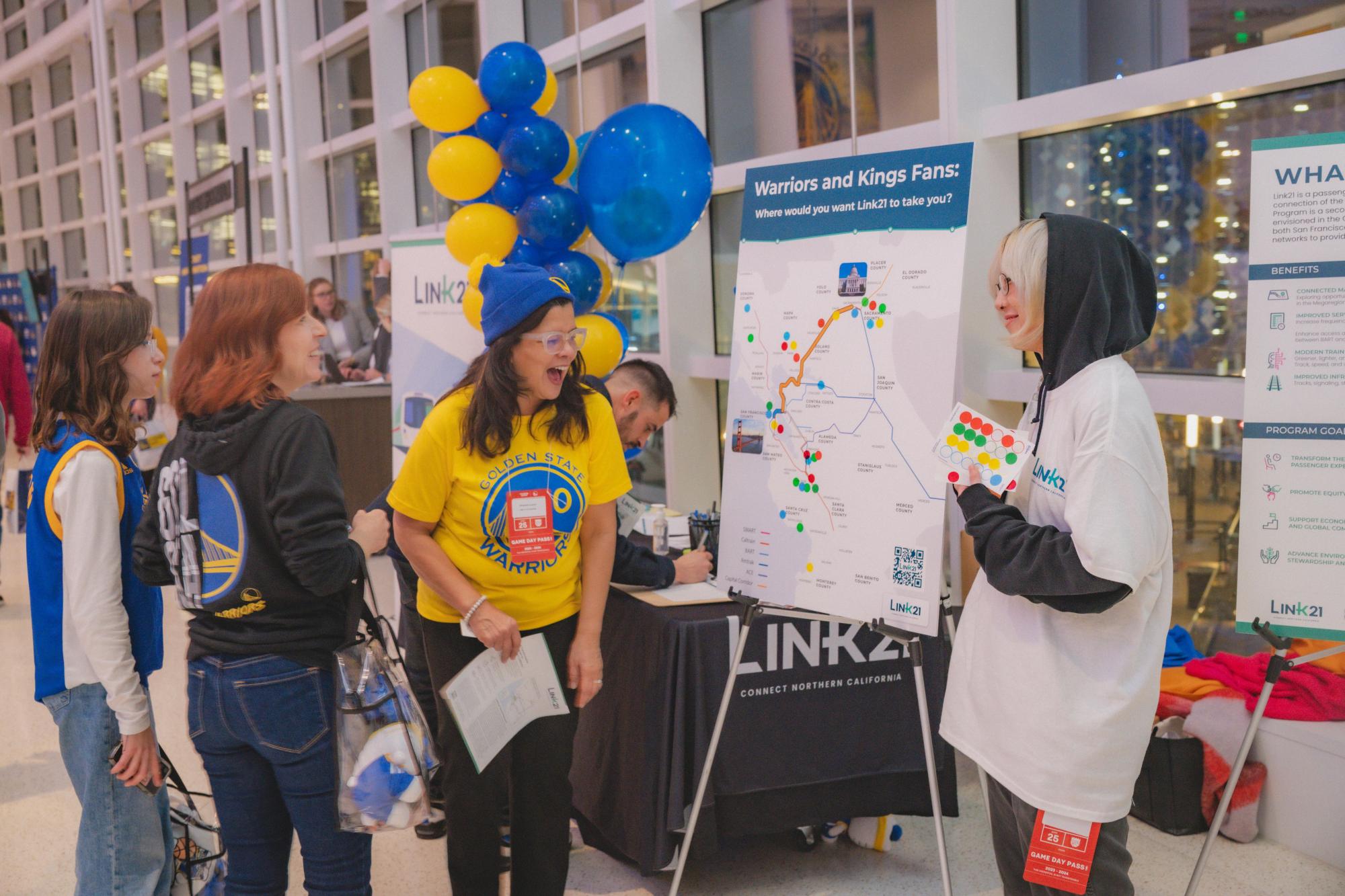 Attendees in Golden State Warriors gear smile in excitement as they interact with staff hosting a Link21 information table.