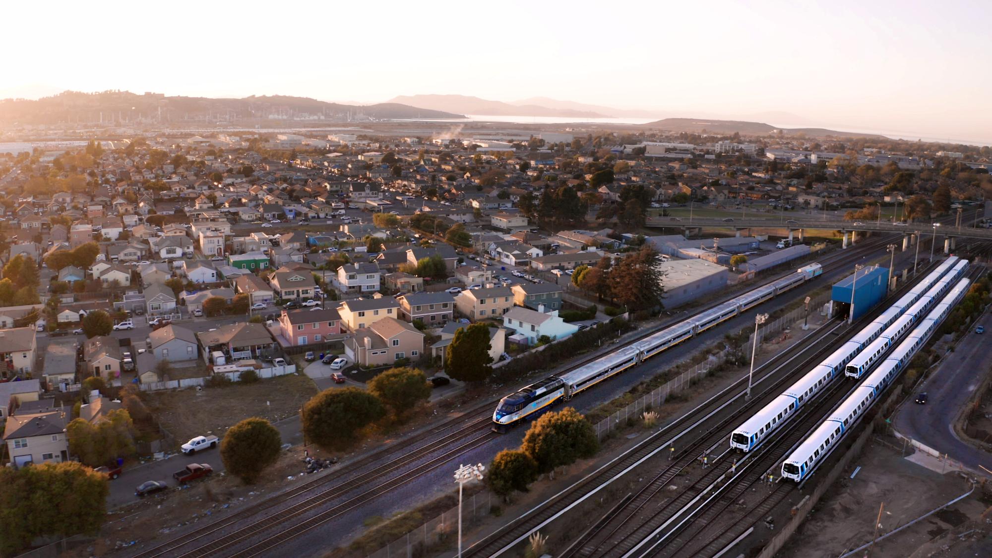 An aerial view in Richmond, California captures a sprawling residential neighborhood adjacent to the right-of-way for Capitol Corridor and BART service with multiple trains on the tracks.