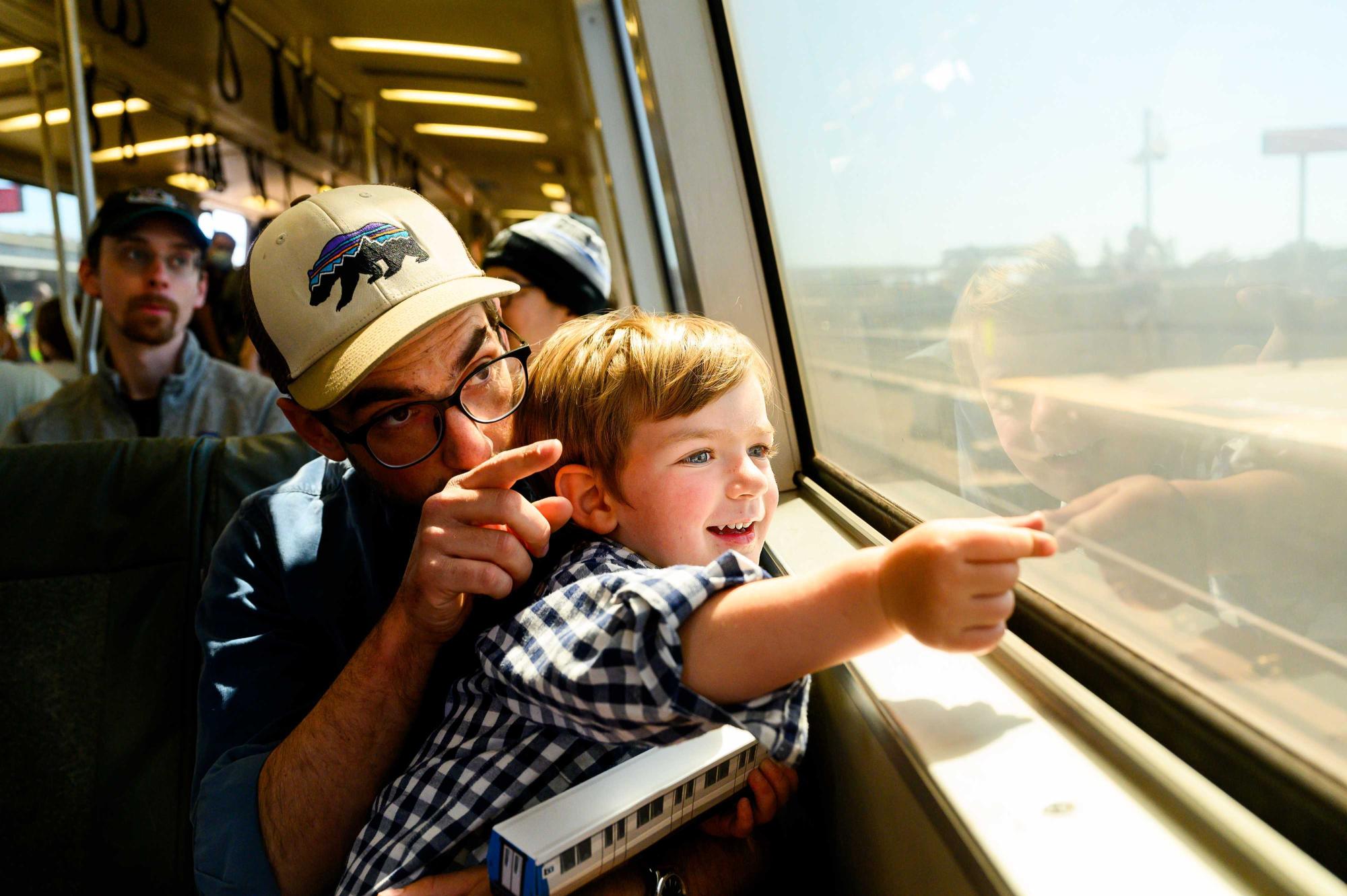 A parent and a child sitting in a BART train both point out the train window with excitement.