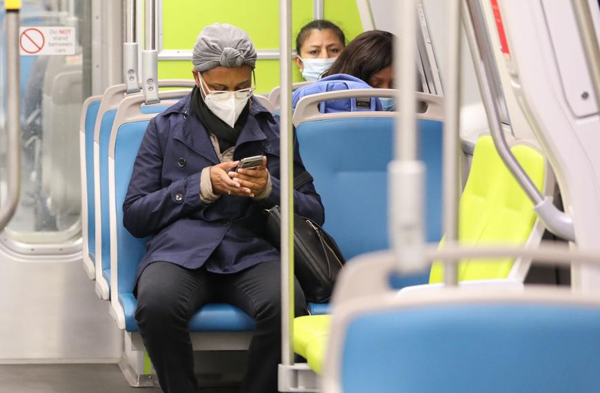 A masked woman on a green and blue Fleet of the Future BART train looks at her phone.
